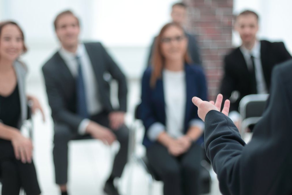 Eine Runde von Führungskräften sitzt im Kreis und hört einer Person zu, die mit offener Hand spricht.